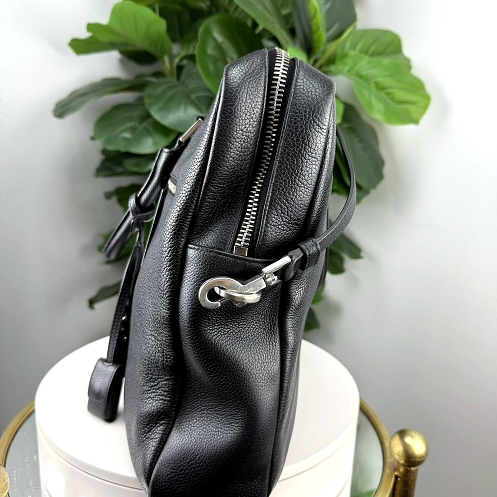 A black pebbled leather crossbody bag with silver-tone hardware, shown standing upright on a white cylindrical pedestal with a green plant in the background.